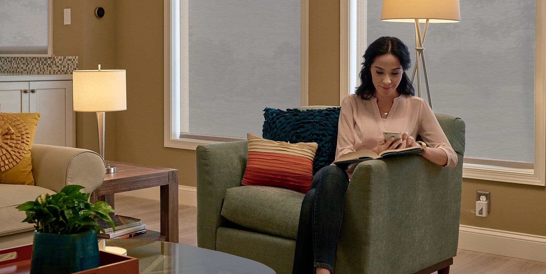 A woman in an armchair using her phone, with roller shades lowered behind her for the evening