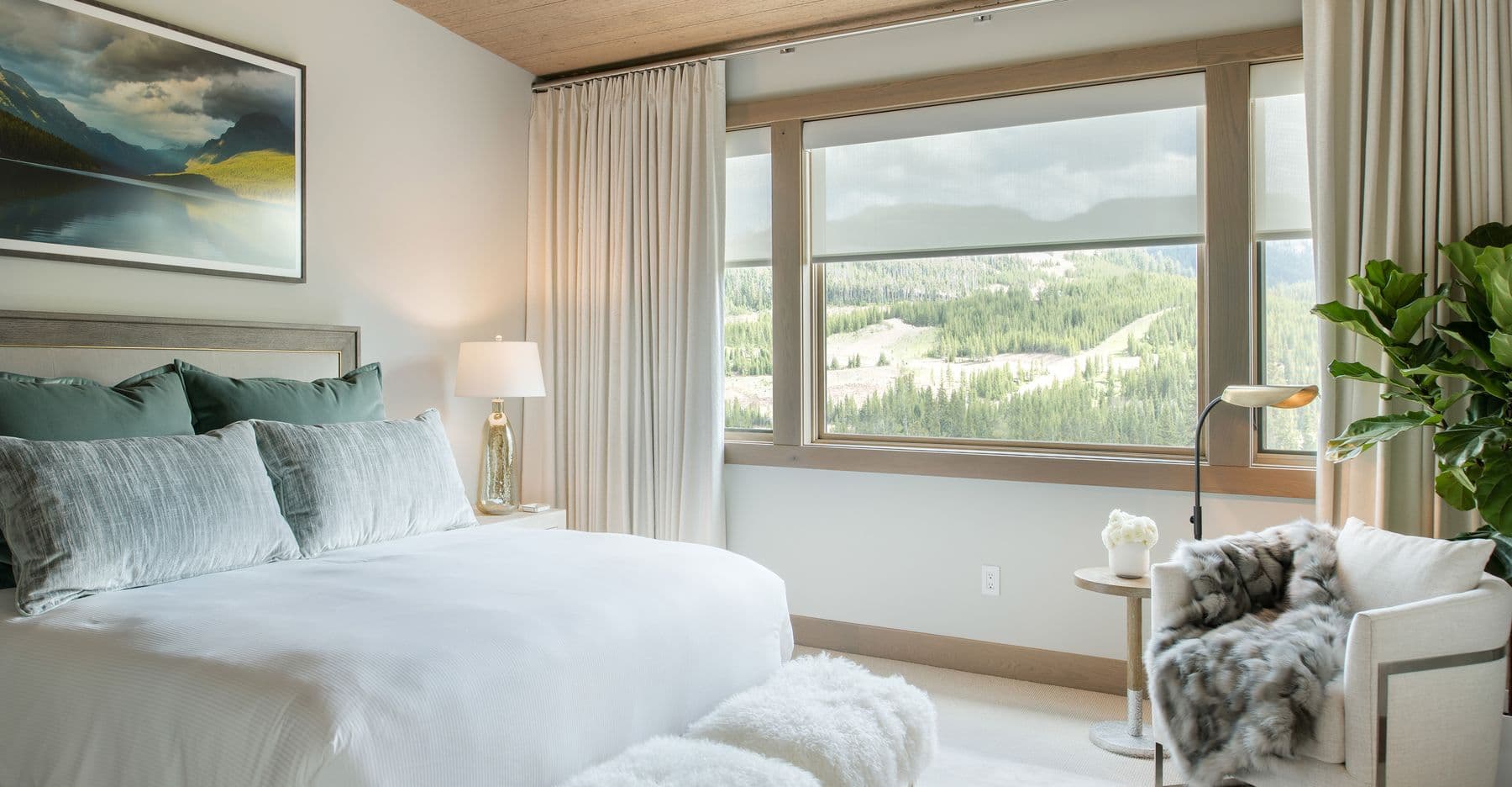 A mountain bedroom with cream drapery on a ceiling track and roller shades partly lowered
