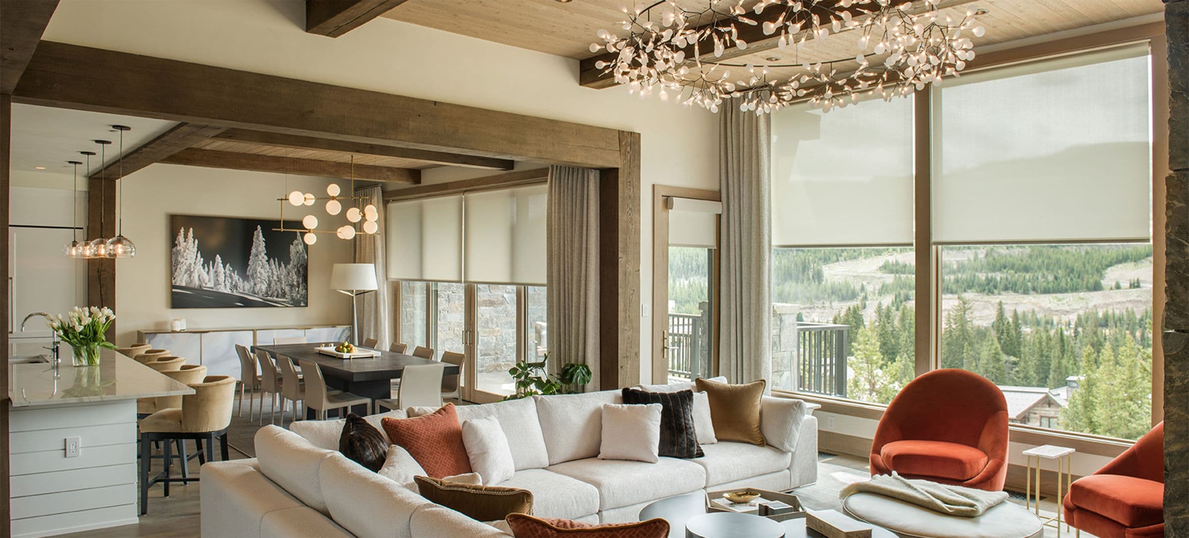 A mountain great room with timber beams, roller shades partly lowered and drapery framing a forest view