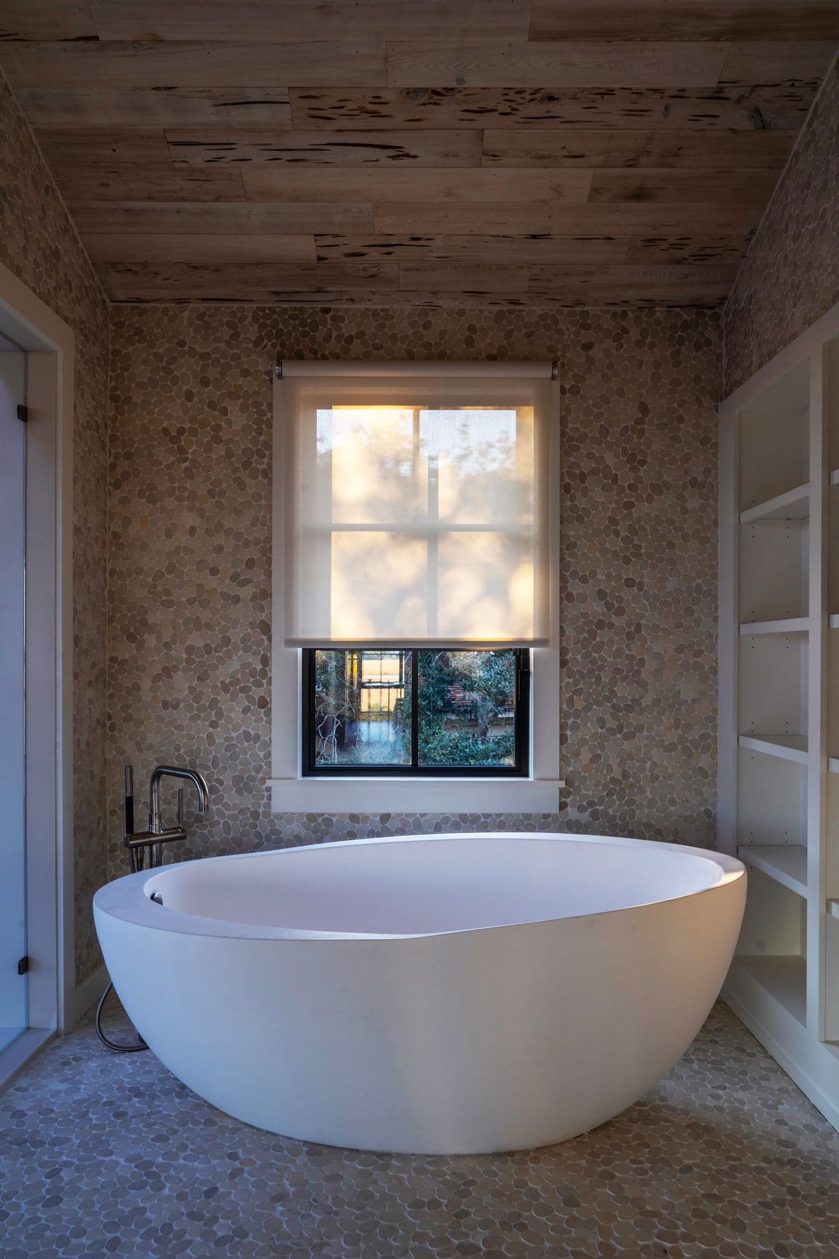 A spa bathroom with a freestanding tub and one sheer shade partly lowered over the window