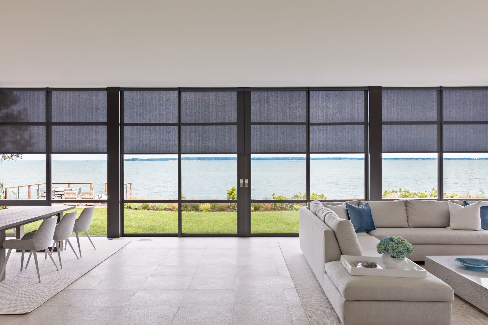 A waterfront living room with a wall of glass and dark screen shades half lowered from ceiling pockets