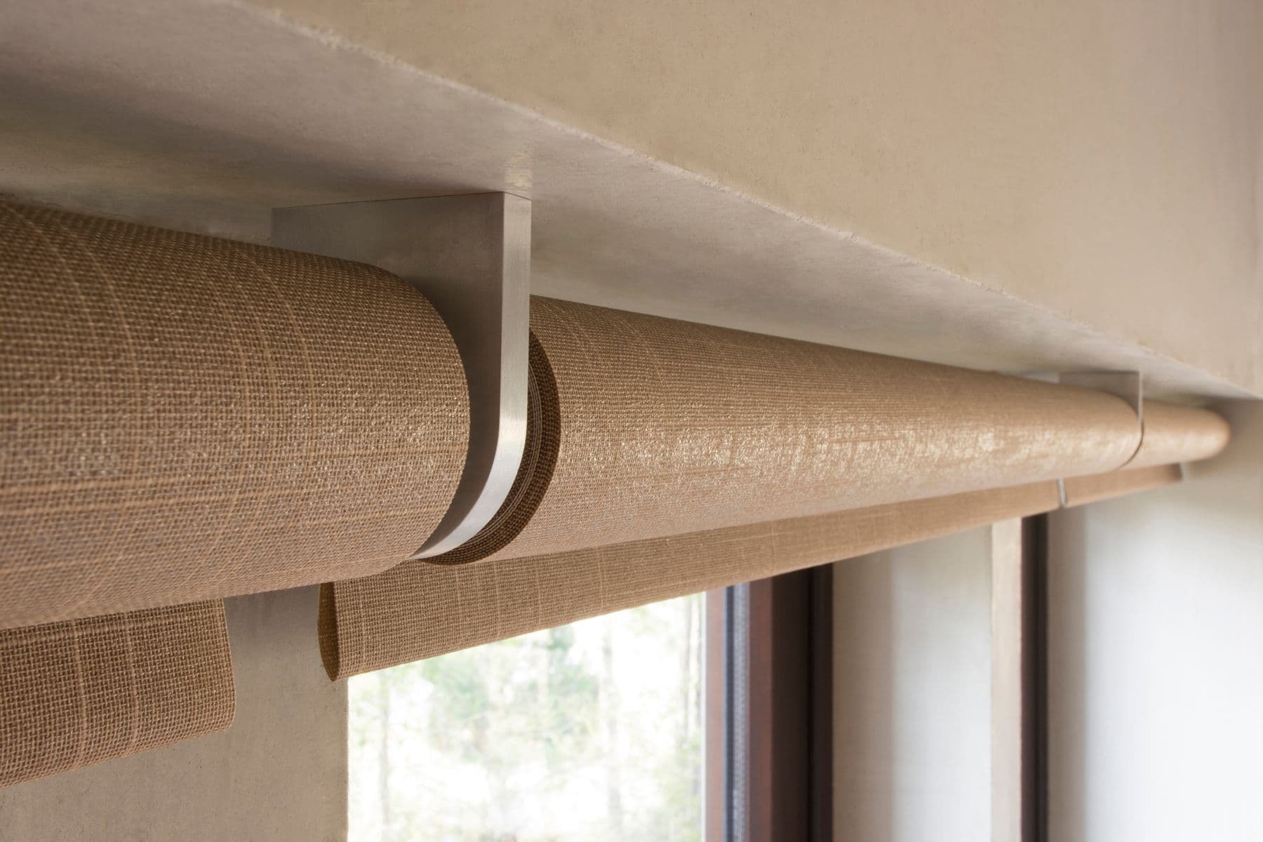 Close-up of woven shade fabric rolled on brushed aluminum brackets under a plaster soffit