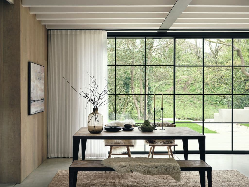 A dining room with a sheer curtain on a motorized track beside a black steel window wall